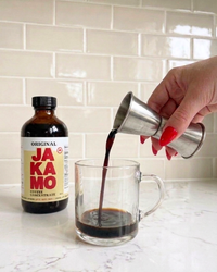 Person pouring coffee concentrate from a jigger into a glass mug with a JAKAMO bottle in the background.