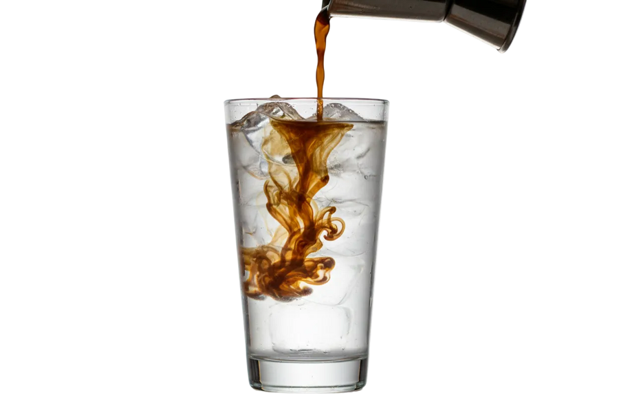 Coffee concentrate being poured from a jigger into a glass of ice water and the coffee is seen spreading through the water like a commercial